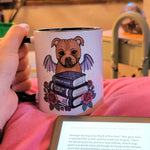 A hand holds a white ceramic mug with a black rim, featuring a cartoon dog with wings and books, in a cozy indoor setting with a tablet in the foreground.