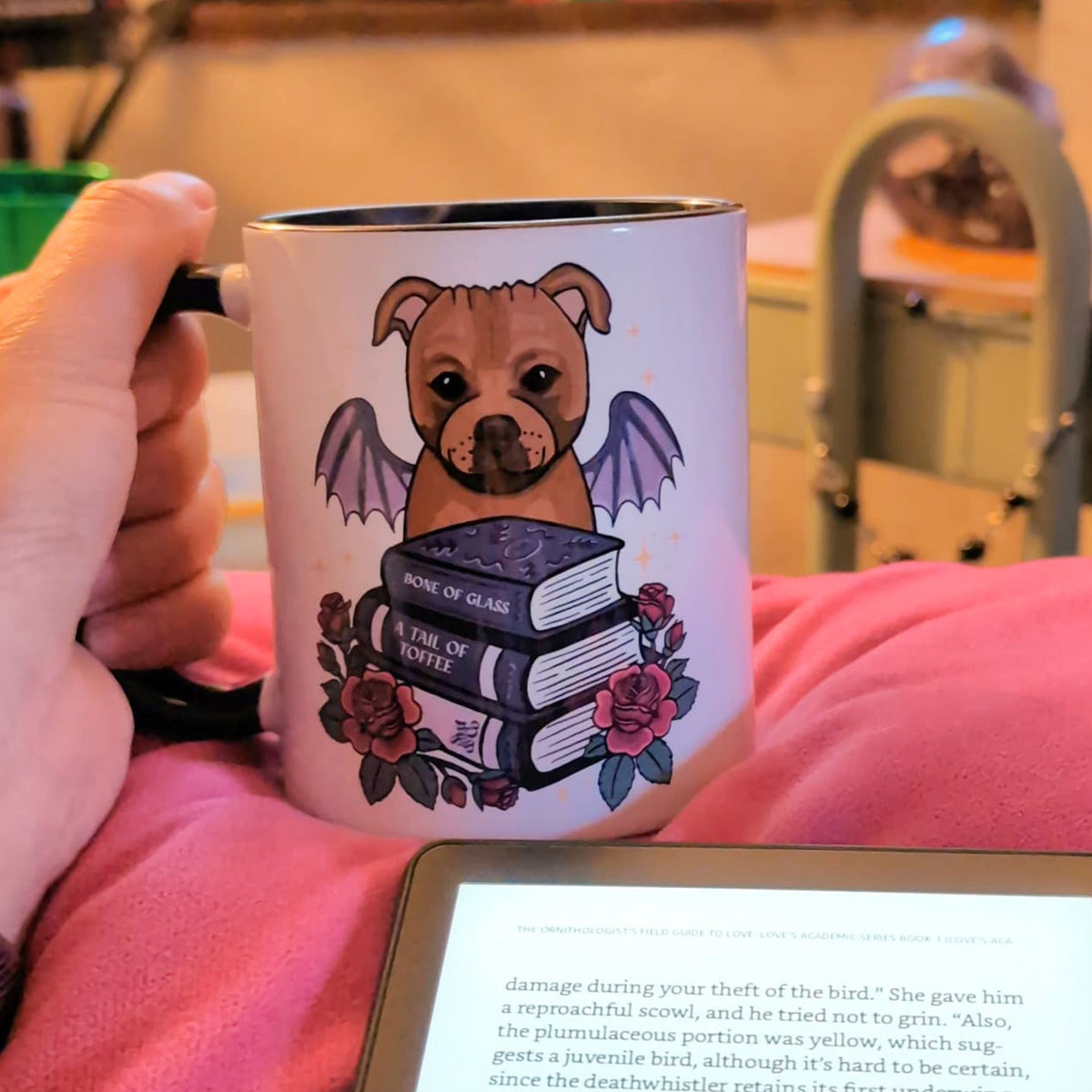 A hand holds a white ceramic mug with a black rim, featuring a cartoon dog with wings and books, in a cozy indoor setting with a tablet in the foreground.