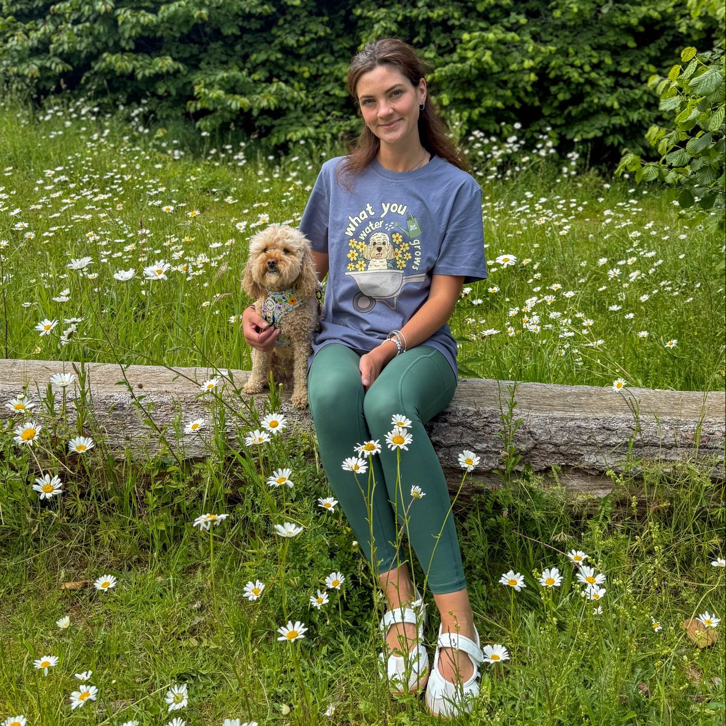 Woman sitting on a log with a dog in a field of flowers and trees