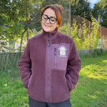 A woman wearing a dusty purple fleece zip-up jacket with a white and green 'The Power of Nature' logo on the chest, standing outdoors on a grassy field with trees and a fence in the background.