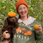 Person wearing an orange beanie and green sweatshirt with a dog and pumpkins, holding small pumpkins in a forest.