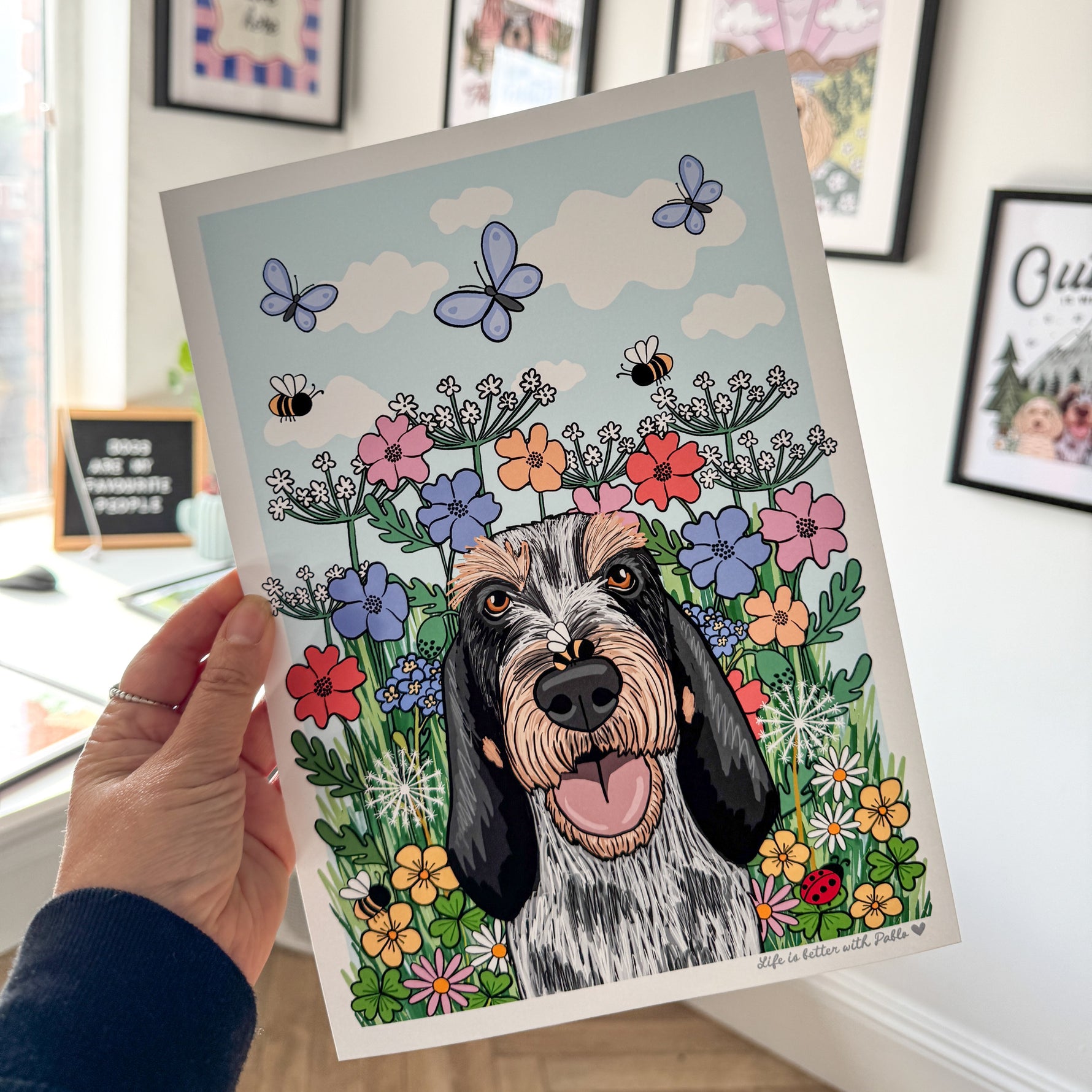 Hand holding a print of a dog surrounded by flowers and butterflies in a room with framed pictures on the wall.