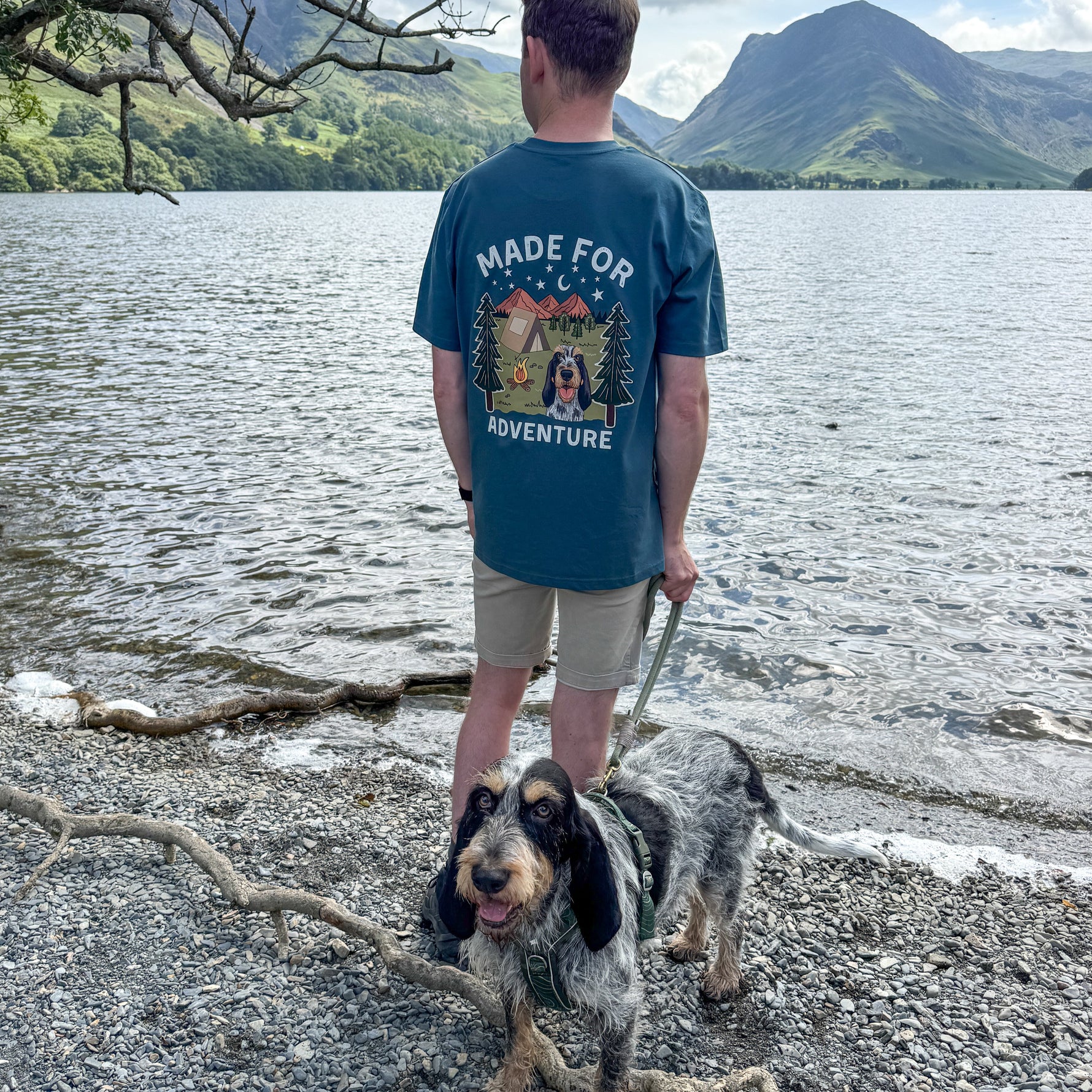 Man walking a dog by a lake with mountains in the background, wearing a t-shirt with a graphic design and text.