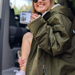 Person in a green jacket holding a cup with a scenic design, sitting inside a vehicle.