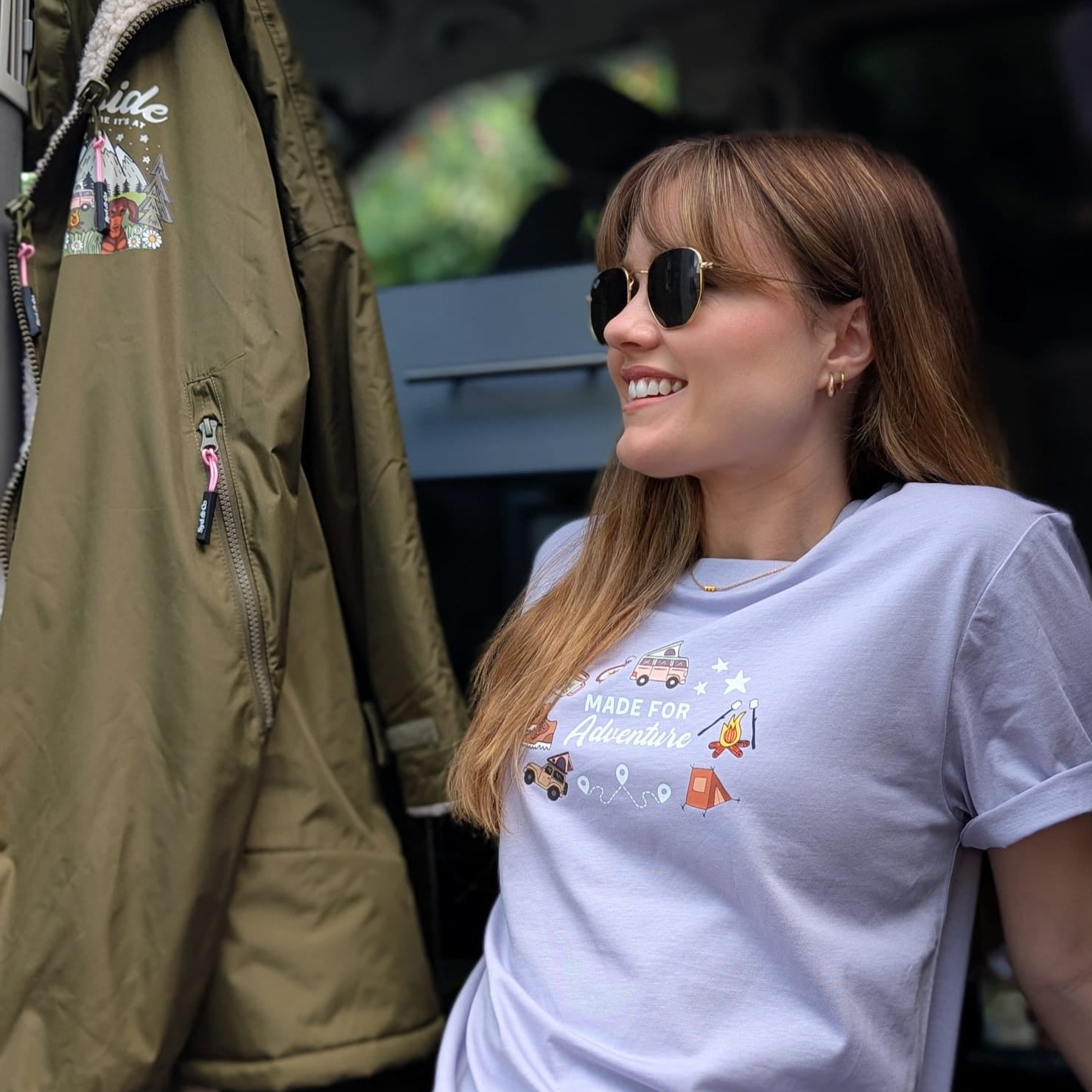 Woman wearing a white t-shirt with graphics and text, sitting next to a green jacket inside a vehicle.