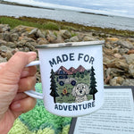 Person holding a mug with 'Made for Adventure' design by a body of water.