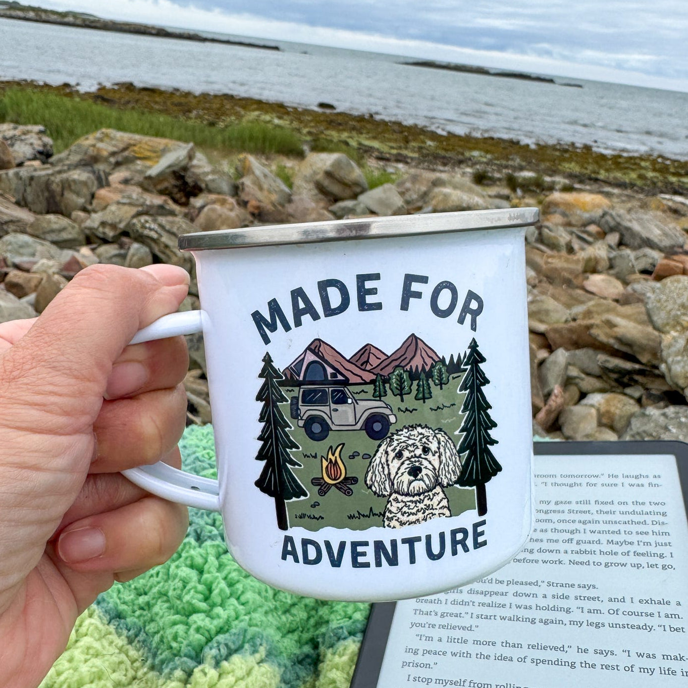 Person holding a mug with 'Made for Adventure' design by a body of water.