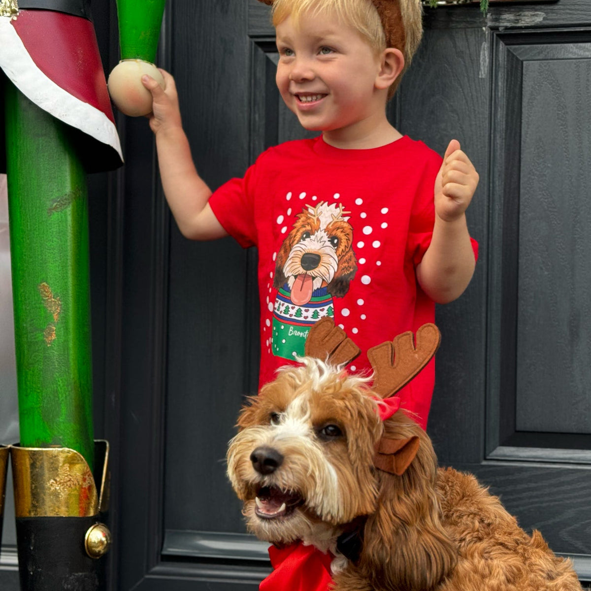 A young child and a dog wearing Christmas-themed headbands and a red holiday shirt with a dog graphic, standing in front of a dark paneled door next to a large nutcracker decoration.