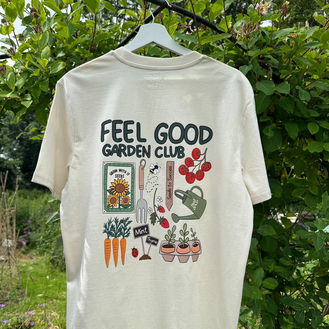 Beige t-shirt with 'Feel Good Garden Club' text and gardening graphics on a hanger against a green garden background.
