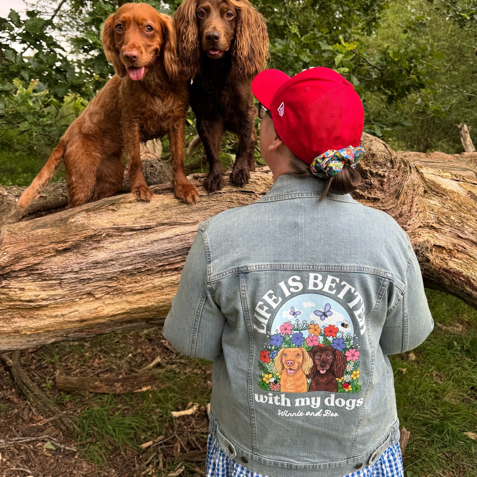 A person wearing a red baseball cap and a denim jacket with a colorful graphic on the back stands on a log outdoors, facing two dogs. The back of the jacket features the text 'LIFE IS BETTER with my dogs' and an illustration of two dogs surrounded by flowers and butterflies.