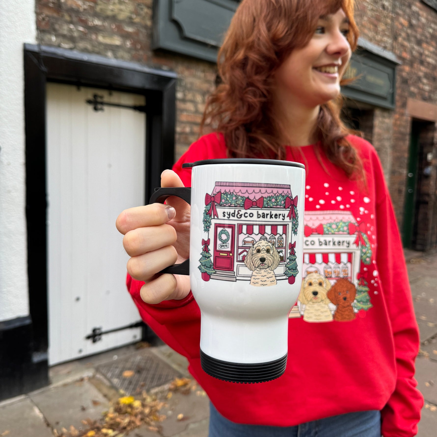 Person wearing a red sweater with a teddy bear design, holding a white mug with a similar design, standing in front of a brick building.