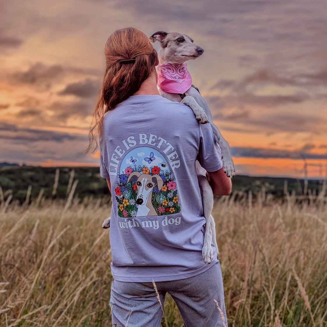 A woman stands in a grassy field at sunset carrying a small dog on her shoulders, wearing a light blue sweatshirt with a large colorful graphic on the back that reads 'LIFE IS BETTER WITH MY DOG.'.