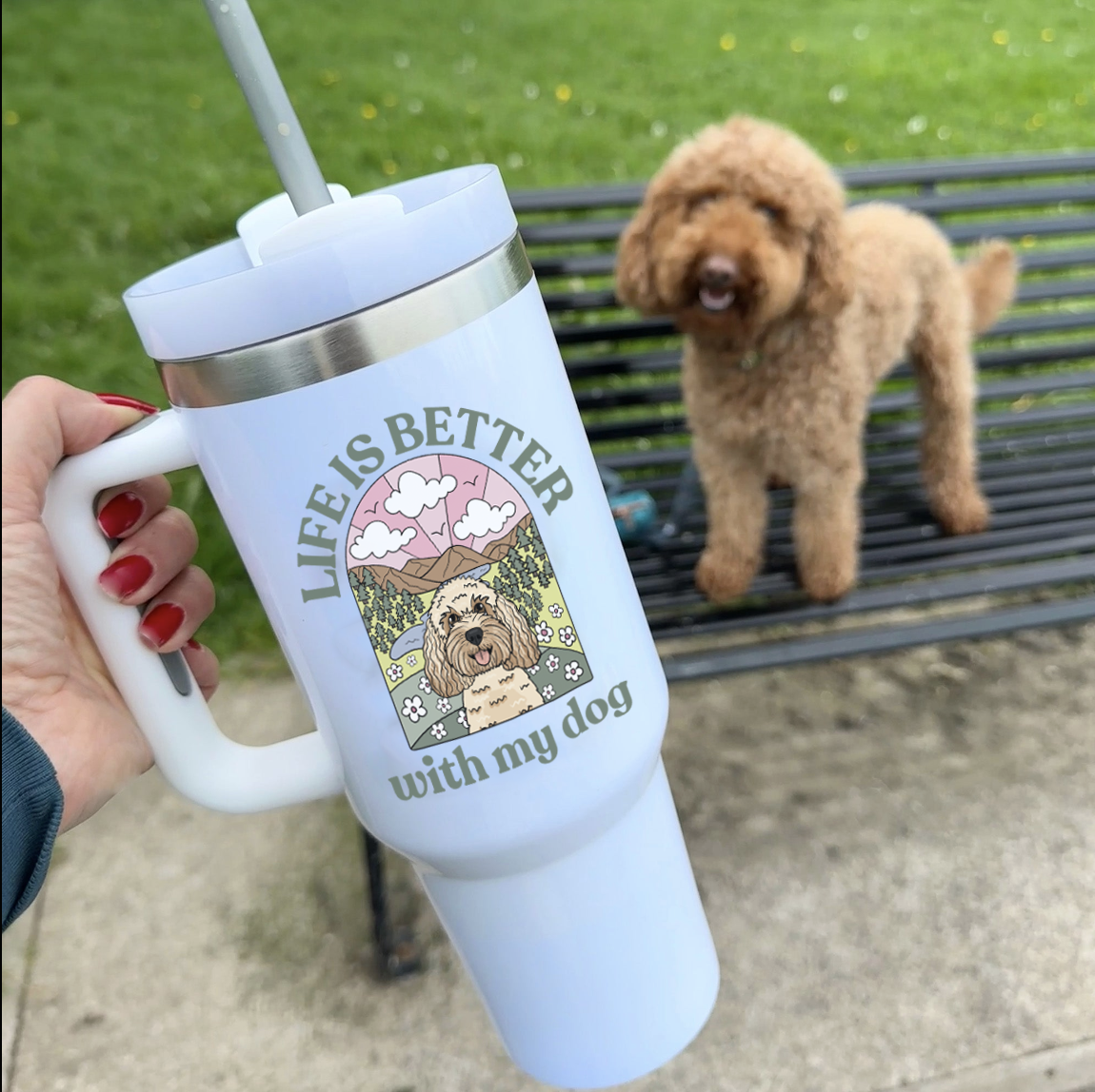 A person holds a light blue insulated tumbler with a straw outdoors, featuring a printed graphic of a dog and the text 'LIFE IS BETTER with my dog.' A fluffy brown dog stands on a bench in the background on a grassy lawn.