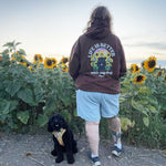 A person standing in a sunflower field with their back to the camera, wearing a brown hoodie with a 'Life is Better with my dog' graphic on the back. A small black dog sits on the ground next to them.