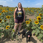 A woman stands in a sunflower field wearing a black graphic vest top that reads 'LIFE IS BETTER with my dog' and a black dog on a leash. The background is filled with rows of sunflowers under a blue sky.