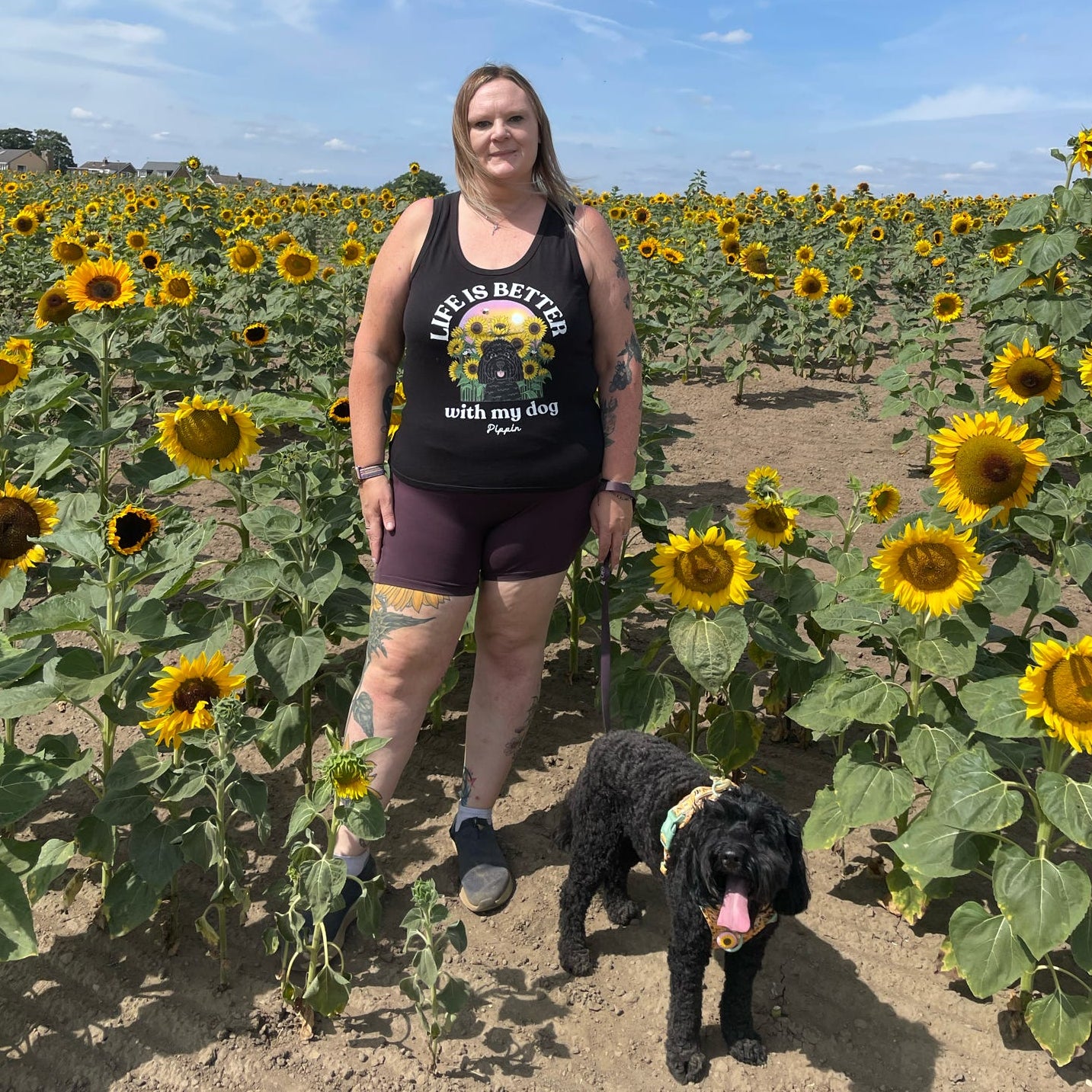 A woman stands in a sunflower field wearing a black graphic vest top that reads 'LIFE IS BETTER with my dog' and a black dog on a leash. The background is filled with rows of sunflowers under a blue sky.