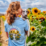 A woman in a light blue graphic t-shirt holds a small white dog in a sunflower field under a blue sky. The back of the shirt features a graphic with sunflowers, a dog, and the text 'LIFE IS BETTER WITH my dog'.