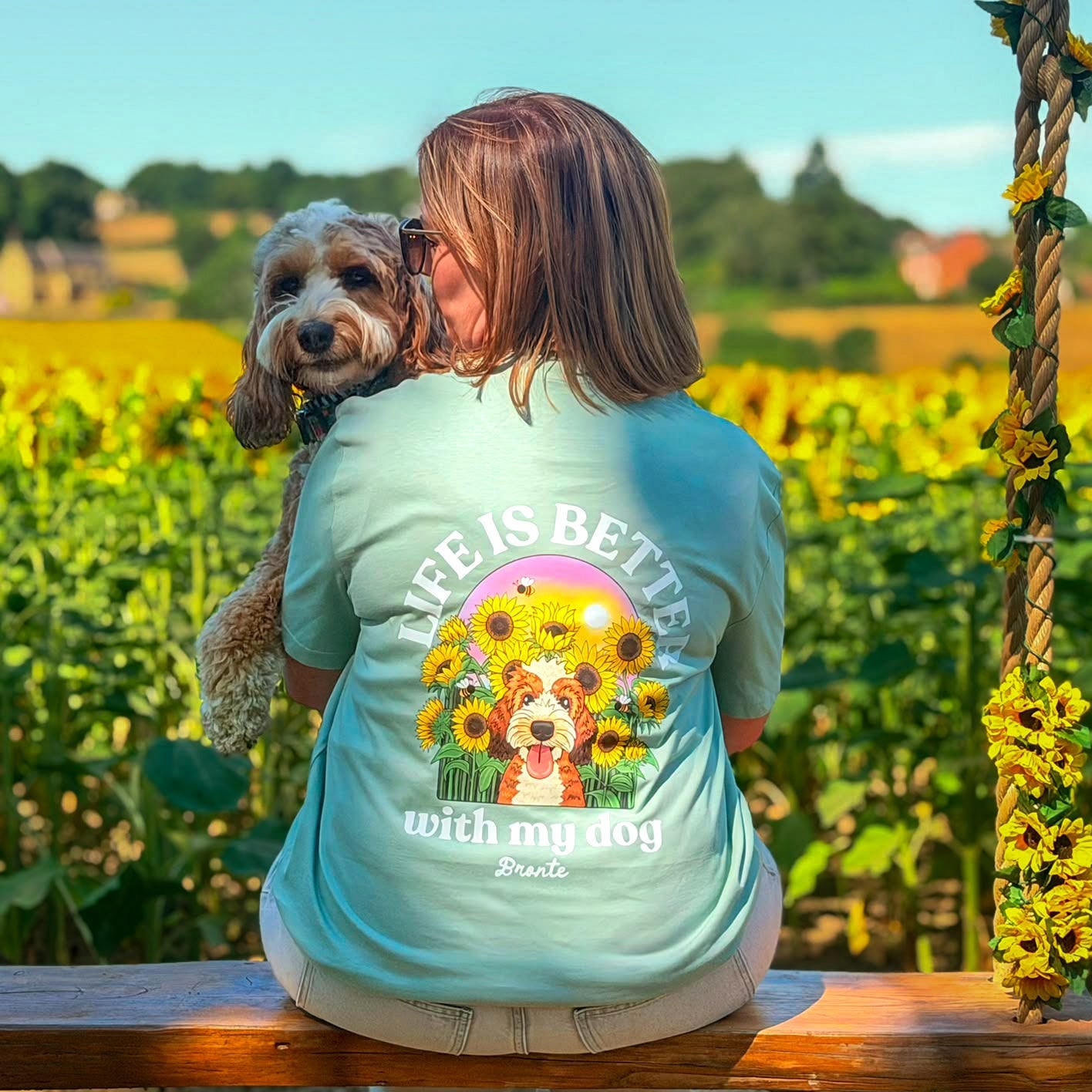 A woman sits on a wooden swing in a sunflower field, holding a small dog. She is wearing a light green graphic t-shirt with a colorful dog illustration and the text 'LIFE IS BETTER WITH MY DOG' on the back.