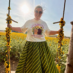 A woman wearing a white graphic t-shirt and a green and yellow striped skirt sits on a rustic wooden swing in a sunflower field.