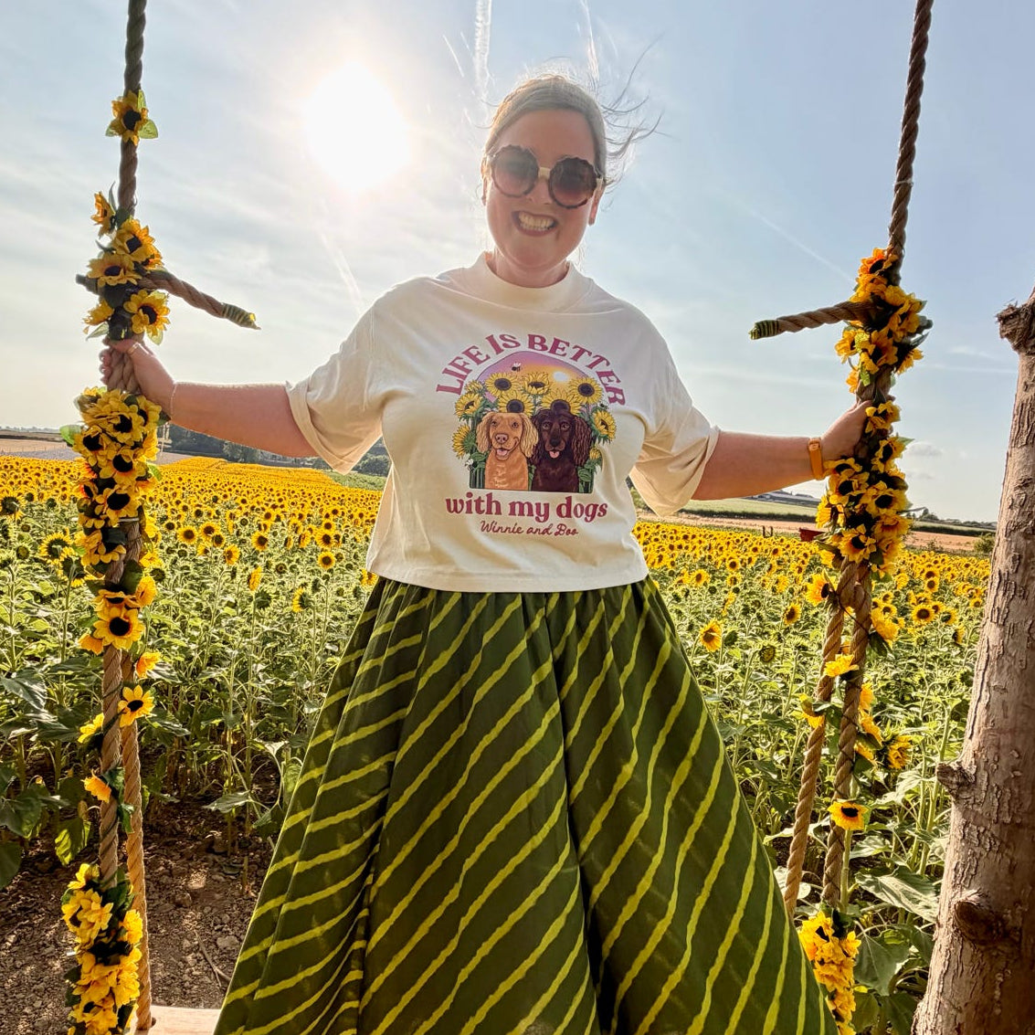 A woman wearing a white graphic t-shirt and a green and yellow striped skirt sits on a rustic wooden swing in a sunflower field.