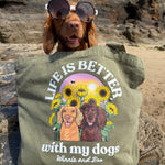 An olive green canvas tote bag with a custom graphic print of two dogs and sunflowers, featuring the text 'LIFE IS BETTER with my dogs Winnie and Boo'. A brown dog wearing sunglasses is sitting behind the bag on a rocky beach background.