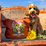 A brown dog sitting on a rustic wooden cart outdoors under a blue sky, next to a brown tote bag printed with 'LIFE IS BETTER with my dog' and a sunflower illustration. A real sunflower rests in front of the dog.