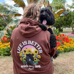 A person sitting outdoors on grass, seen from behind, wearing a brown hoodie with a large graphic on the back that reads 'LIFE IS BETTER with my dog' around a sunflower and dog illustration. A black dog is sitting on the person's shoulder in a garden setting with flowers and banana leaves in the background.