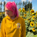 A person with bright pink hair wearing a mustard yellow hoodie stands outdoors in front of a field of sunflowers under a blue sky. The hoodie has a small  graphic on the left chest that reads 'Life is Better with my dogs'.