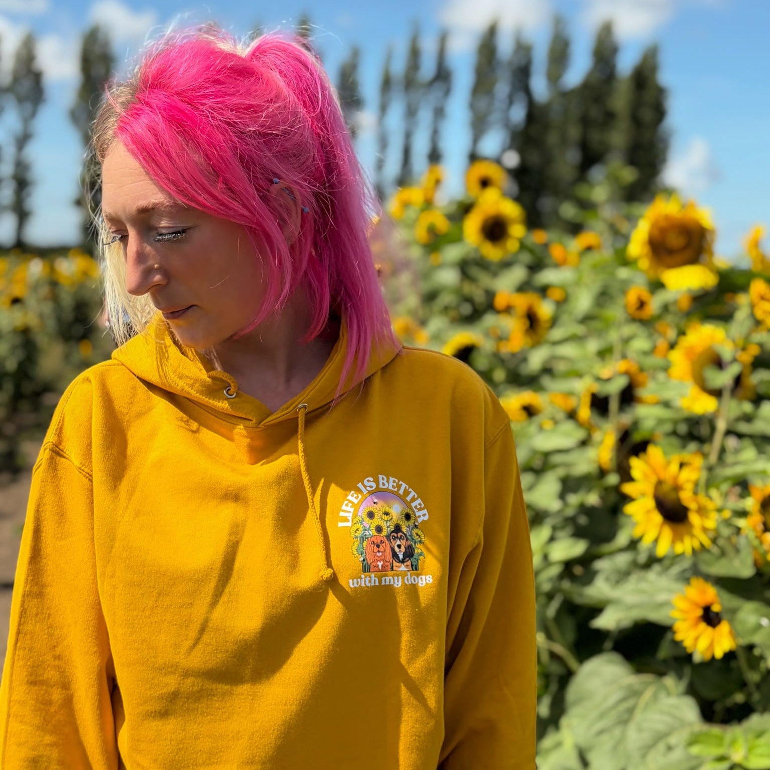 A person with bright pink hair wearing a mustard yellow hoodie stands outdoors in front of a field of sunflowers under a blue sky. The hoodie has a small  graphic on the left chest that reads 'Life is Better with my dogs'.
