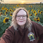 A woman wearing glasses and a brown hoodie with a sunflower-themed graphic on the chest, posing for a selfie in a sunflower field at sunset.