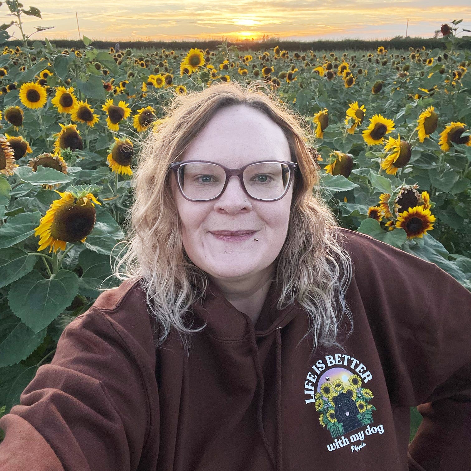 A woman wearing glasses and a brown hoodie with a sunflower-themed graphic on the chest, posing for a selfie in a sunflower field at sunset.