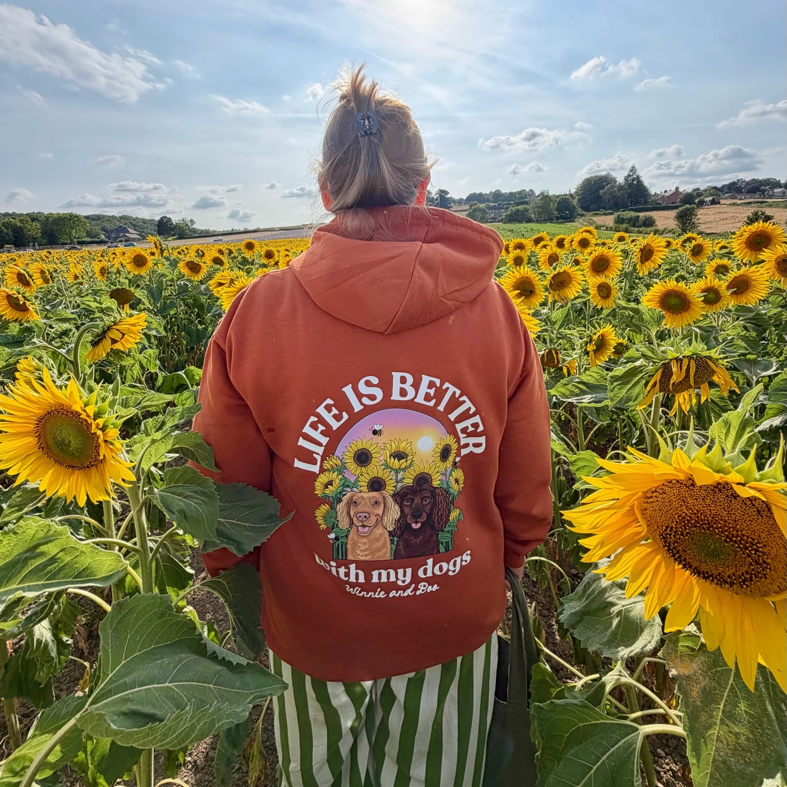 A person standing in a sunflower field, viewed from behind, wearing a rust-orange hoodie with a large graphic on the back that reads 'LIFE IS BETTER WITH MY DOGS' and a photo of two dogs.