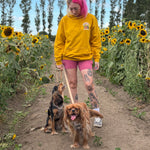 A woman with pink hair walks two dogs on a dirt path between rows of sunflowers, wearing a yellow hoodie, pink shorts, and white sneakers.