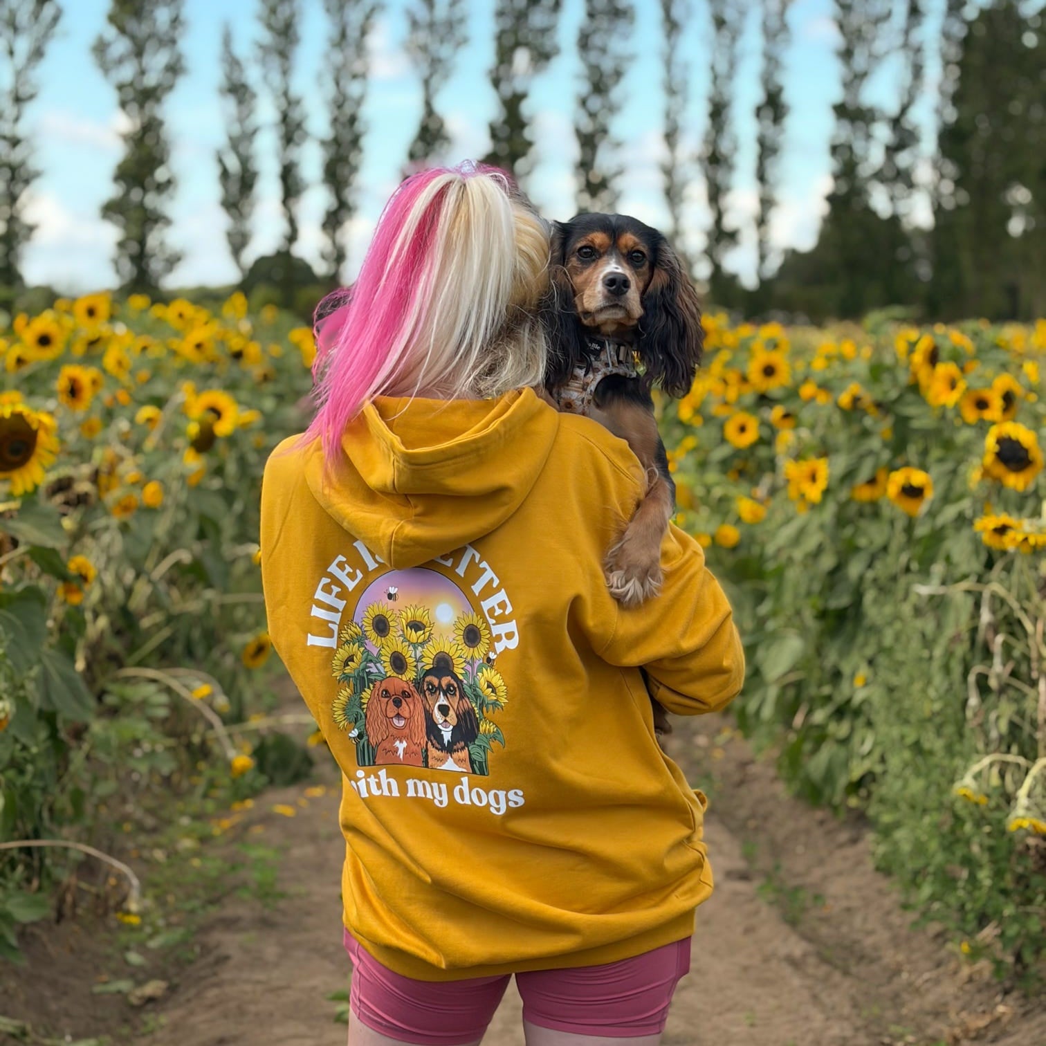 A woman standing in a sunflower field holding a small black and tan dog, viewed from behind. She is wearing a bright yellow hoodie with a large graphic on the back that reads 'LIFE IS BETTER WITH MY DOGS' and shows two dogs in a garden scene.