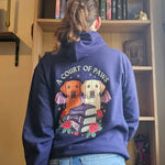 A person viewed from behind wearing a navy blue hoodie with a large graphic on the back. The graphic features the text 'A COURT OF PAWS' above two dogs with bat wings above a stack of books and roses.