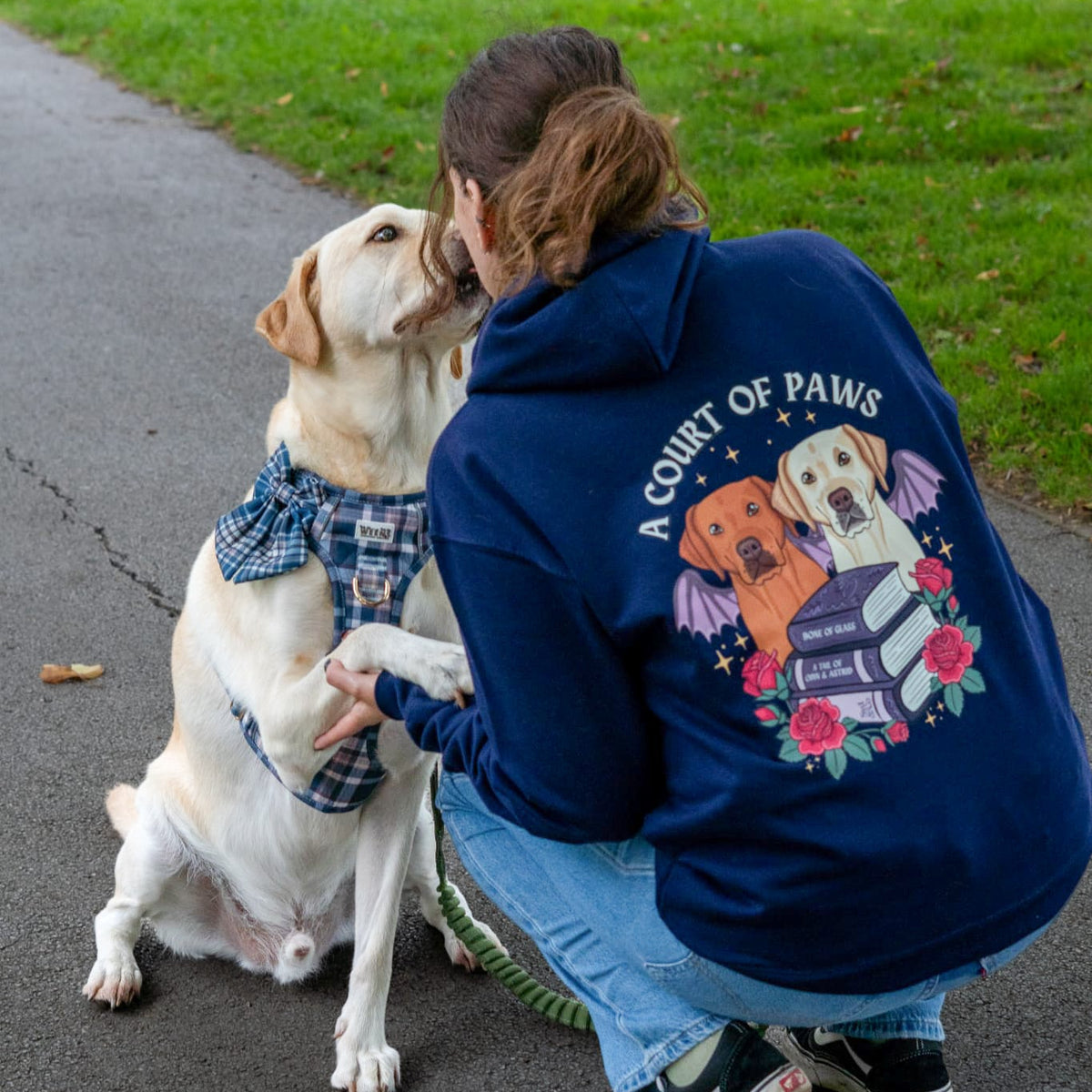 A woman crouches outdoors on a paved path next to a grassy lawn, wearing a navy blue hoodie with a large graphic on the back featuring two dogs and the text 'A COURT OF PAWS'. She is interacting with a light-colored dog wearing a matching plaid harness and collar.