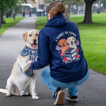 A woman crouches beside a light-colored Labrador retriever on a paved path in a park. The woman wears a navy blue hoodie with a large graphic on the back featuring two dogs, roses, and the text 'A COURT OF PAWS', paired with blue jeans.