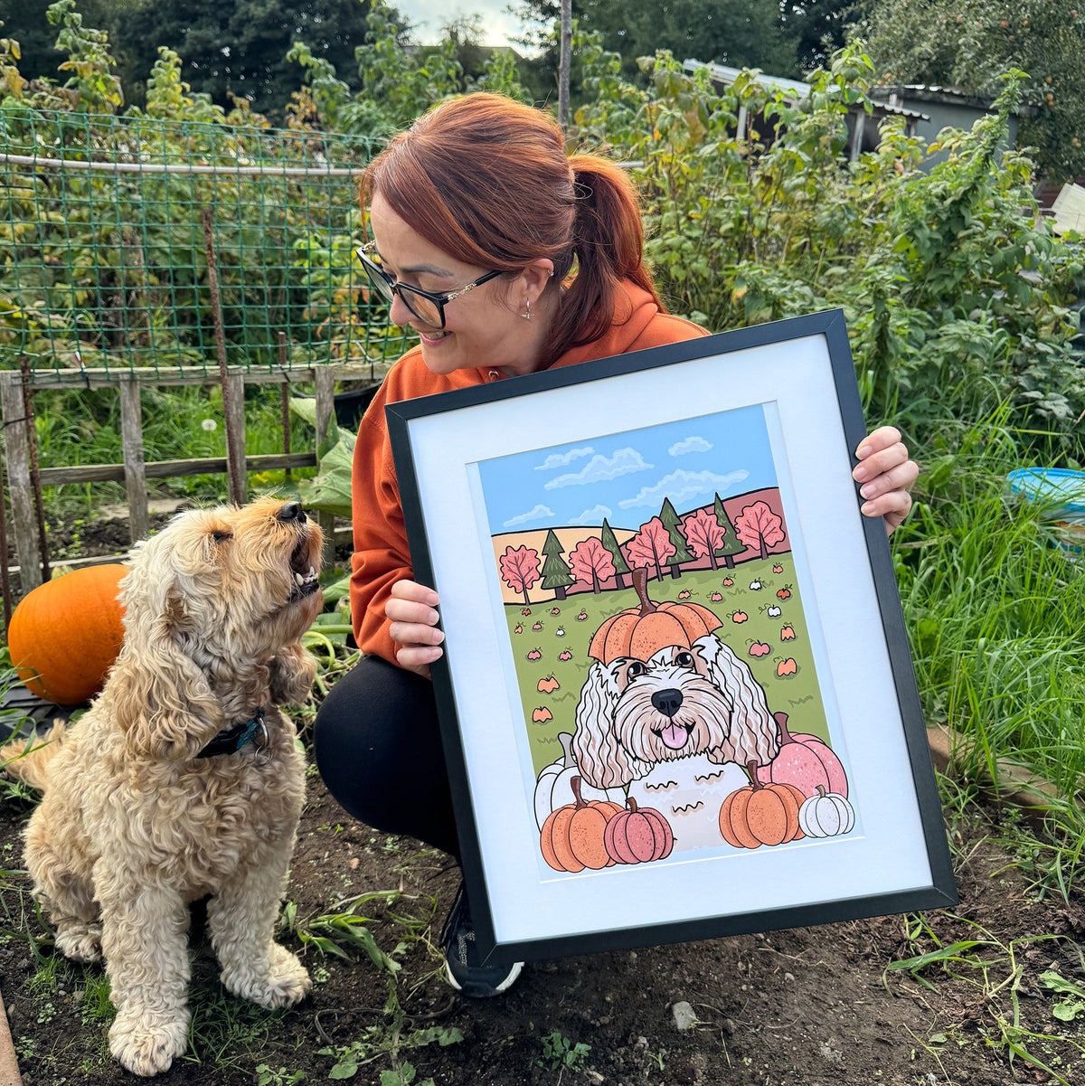 A woman in an orange hoodie and glasses holds up a framed custom pet portrait in a garden setting, next to a fluffy golden dog. The portrait features a cartoon-style drawing of a dog with a pumpkin on its head, surrounded by pumpkins and trees under a blue sky.