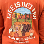 An orange hoodie shown from the back, worn by a person outdoors. The hoodie features a large graphic print with the text 'LIFE IS BETTER with my pupkins' and an illustration of two dogs sitting in a pumpkin patch.