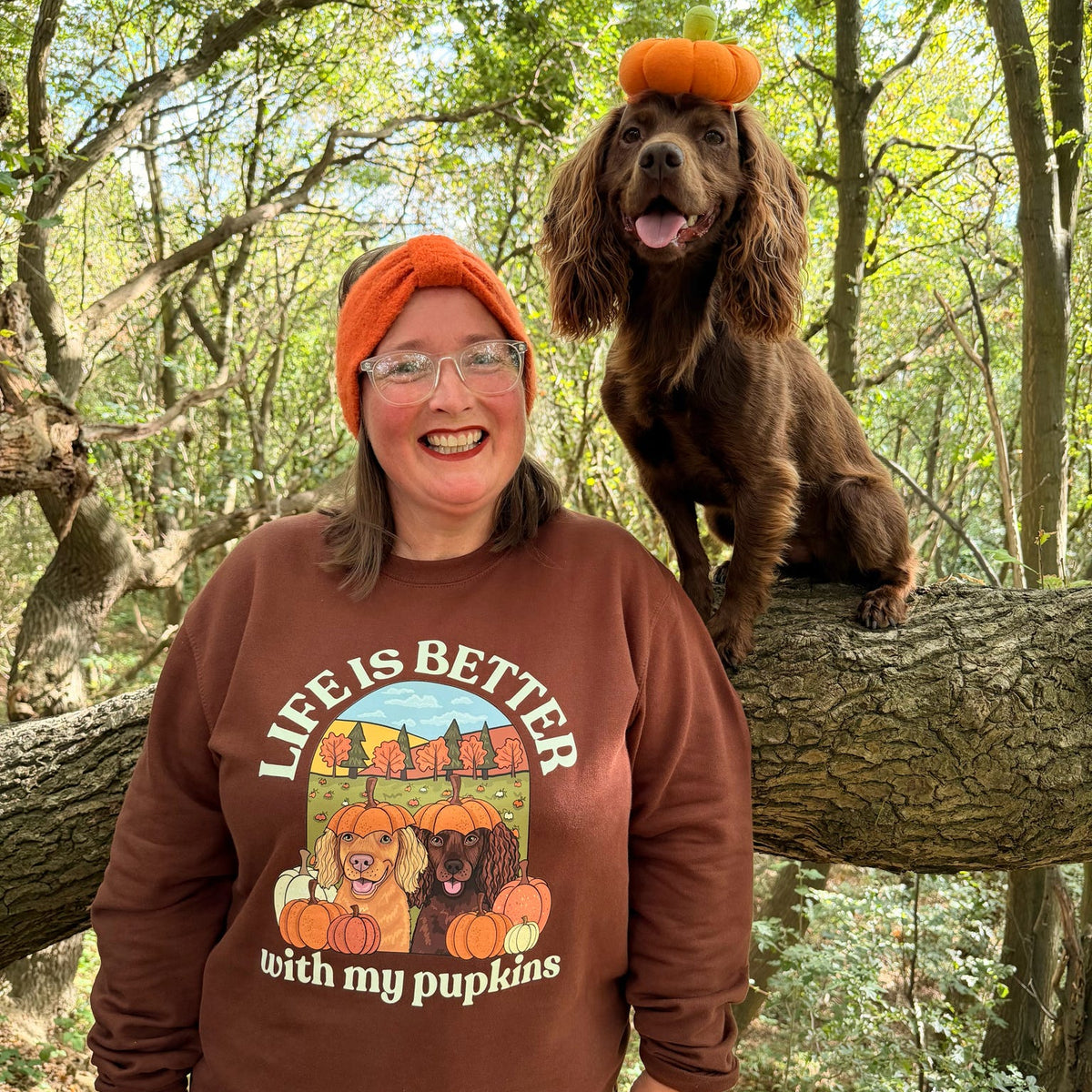 A woman wearing a brown sweatshirt with a 'Life is Better with my pumpkins' graphic and an orange beanie stands next to a brown dog wearing an orange pumpkin topper in a wooded outdoor setting.