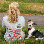 A woman and her dog sit on a grassy hill outdoors. The woman is seen from behind wearing a light gray graphic T-shirt with autumn themed illustrations and the text 'FEEL GOOD WALKING CLUB