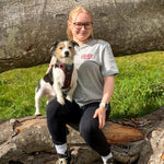 A woman wearing a grey autumn outdoor inspired t-shirt with a logo sits outdoors on a log with a small black, white, and tan dog on her lap. The background features a large fallen tree trunk and green grass.