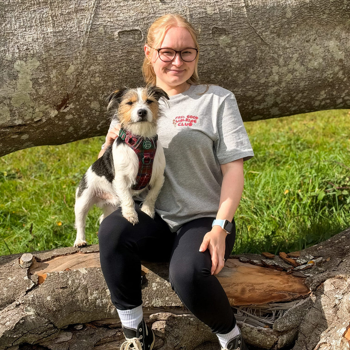 A woman wearing a grey autumn outdoor inspired t-shirt with a logo sits outdoors on a log with a small black, white, and tan dog on her lap. The background features a large fallen tree trunk and green grass.