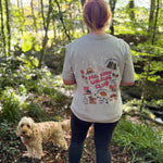 A woman standing in a wooded outdoor setting with her dog, viewed from behind. She is wearing a light heather grey t-shirt with a colorful autumn inspired graphic on the back that reads 'FEEL GOOD WALKING CLUB' surrounded by woodland-themed illustrations.