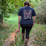 A woman walking a tan dog on a leaf-covered outdoor path, viewed from behind. The woman wears a navy autumn inspired graphic T-shirt, black leggings, and brown-and-tan striped knee-high socks.
