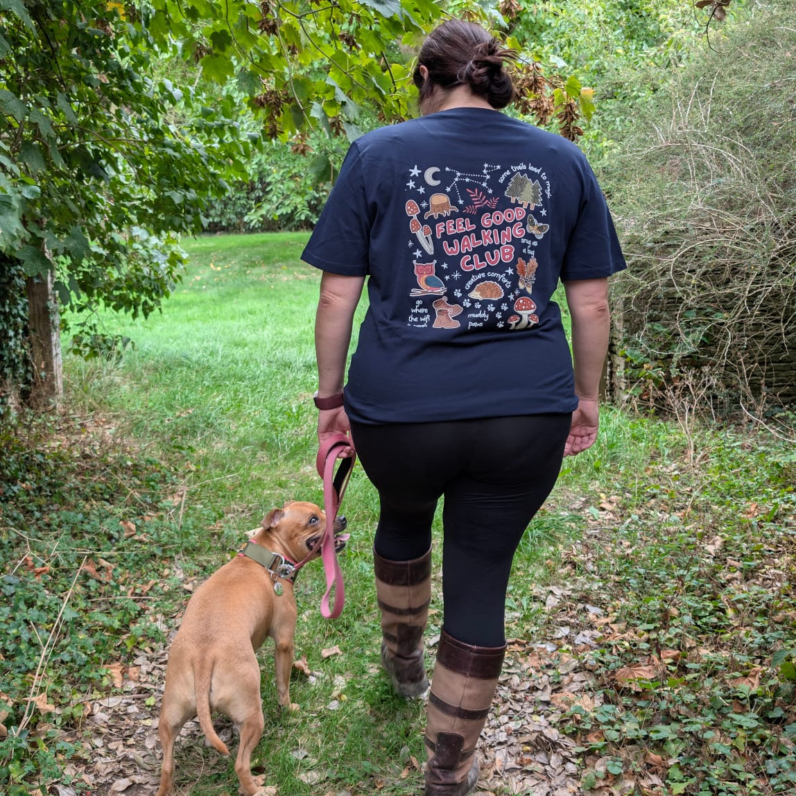 A woman walking a tan dog on a leaf-covered outdoor path, viewed from behind. The woman wears a navy autumn inspired graphic T-shirt, black leggings, and brown-and-tan striped knee-high socks.