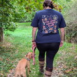 A woman seen from behind walking a tan dog on a leaf-covered outdoor path. The woman is wearing a navy blue graphic T-shirt with a colorful autmun outdoor -style design and the words 'Feel Good Walking Club' across the back, along with black leggings and brown-and-tan knee-high boots.