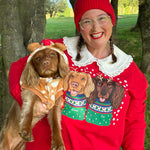 A woman outdoors wearing a bright red Christmas sweater with a white lace collar and a graphic of two dogs in holiday sweaters, holding a brown dog dressed in a dog costume. She is wearing a matching red knit beanie.