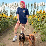 A woman with pink hair stands in a sunflower field holding leashes for two dogs. She is wearing a navy blue graphic t-shirt, pink shorts, and sneakers.