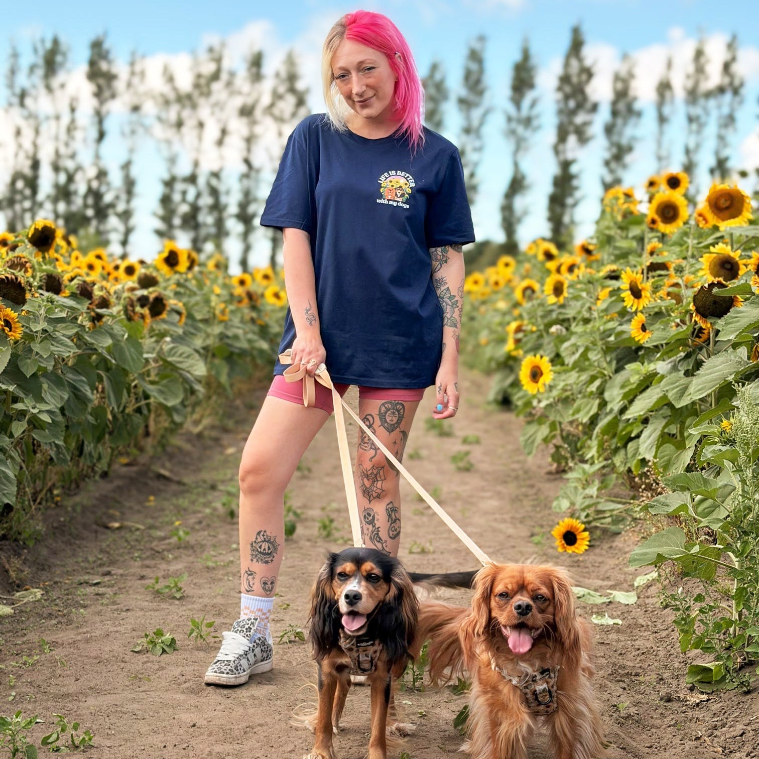 A woman with pink hair stands in a sunflower field holding leashes for two dogs. She is wearing a navy blue graphic t-shirt, pink shorts, and sneakers.